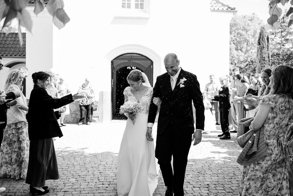 Bride and groom walk down cobblestone path after church wedding ceremony while guests celebrate.