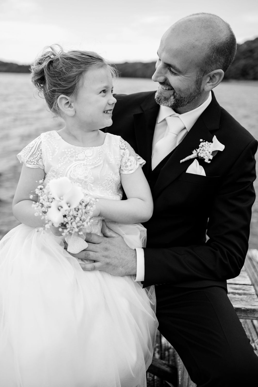 Groom in black tuxedo smiling at flower girl in white dress holding bouquet on dock, black and white wedding photo.