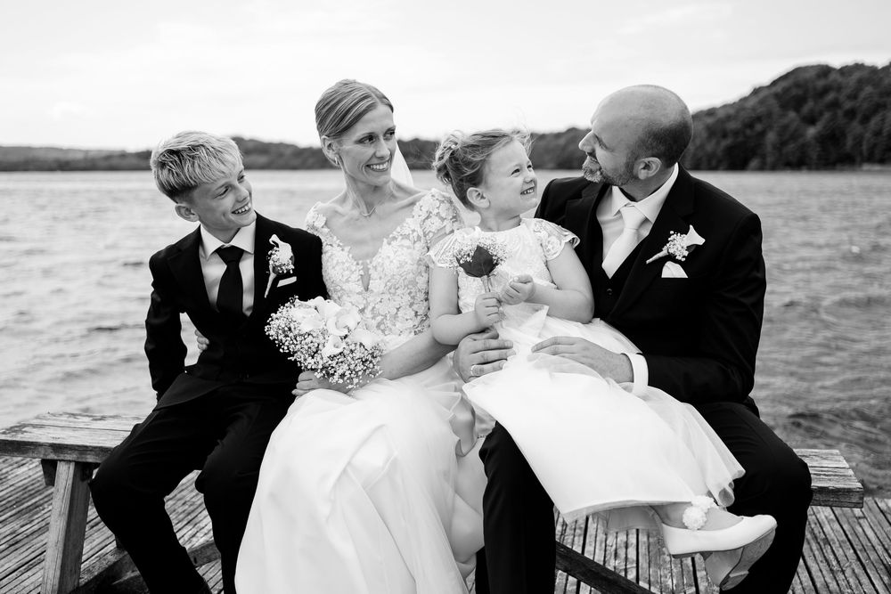 Black and white wedding photo of bride and groom sitting on dock with two children, lake in background.