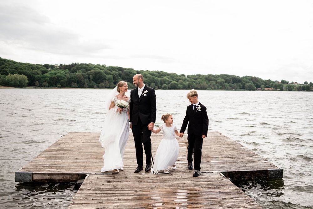 Bride and groom stand on a lakeside dock with two children on a cloudy wedding day.