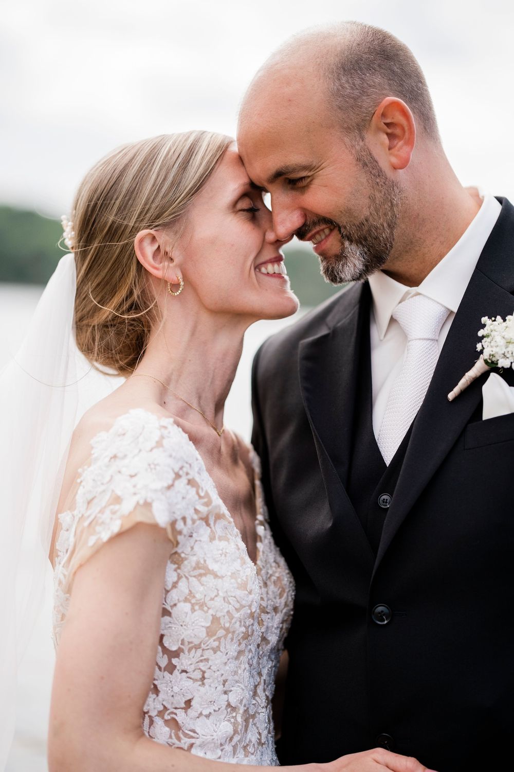 Smiling bride and groom touching foreheads intimately on their wedding day near a lake.