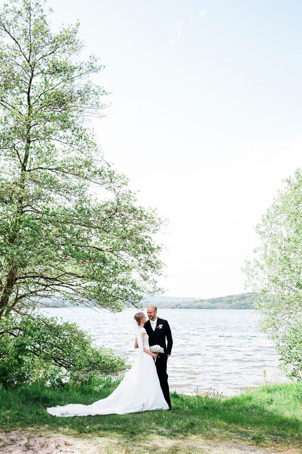 Bride and groom share romantic moment by a serene lake surrounded by lush green trees.