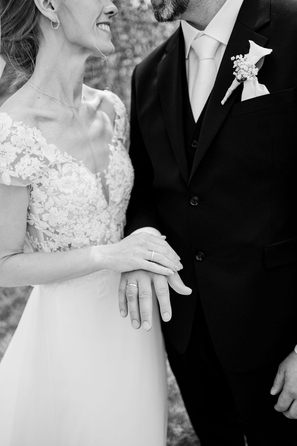 Black and white photo of bride and groom showing wedding rings, bride in lace dress, groom in dark suit with boutonniere.
