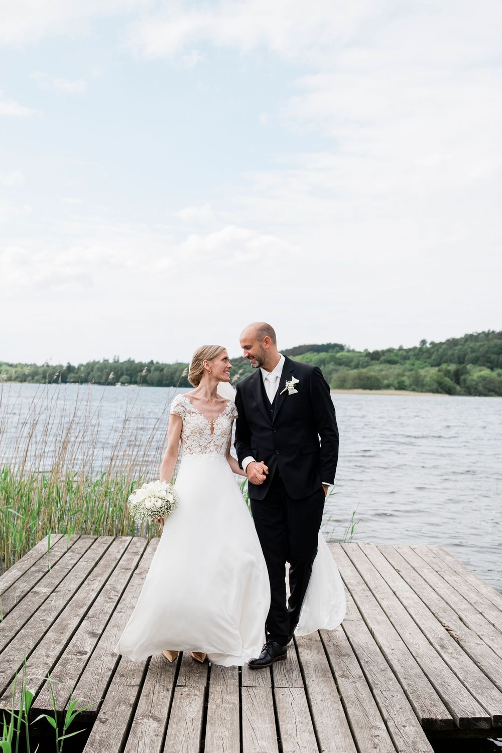 Bride and groom share romantic moment on wooden dock by lake, bride holding white bouquet.