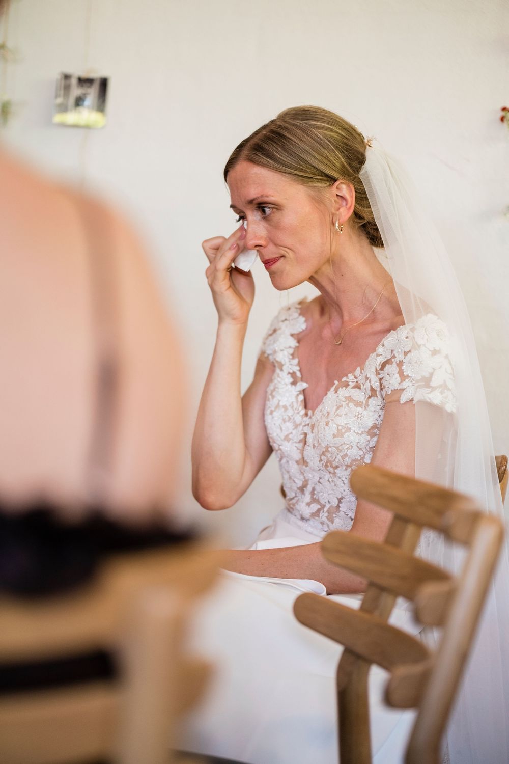Emotional bride in lace wedding dress and veil wiping tears while seated on wooden chair during wedding preparations.