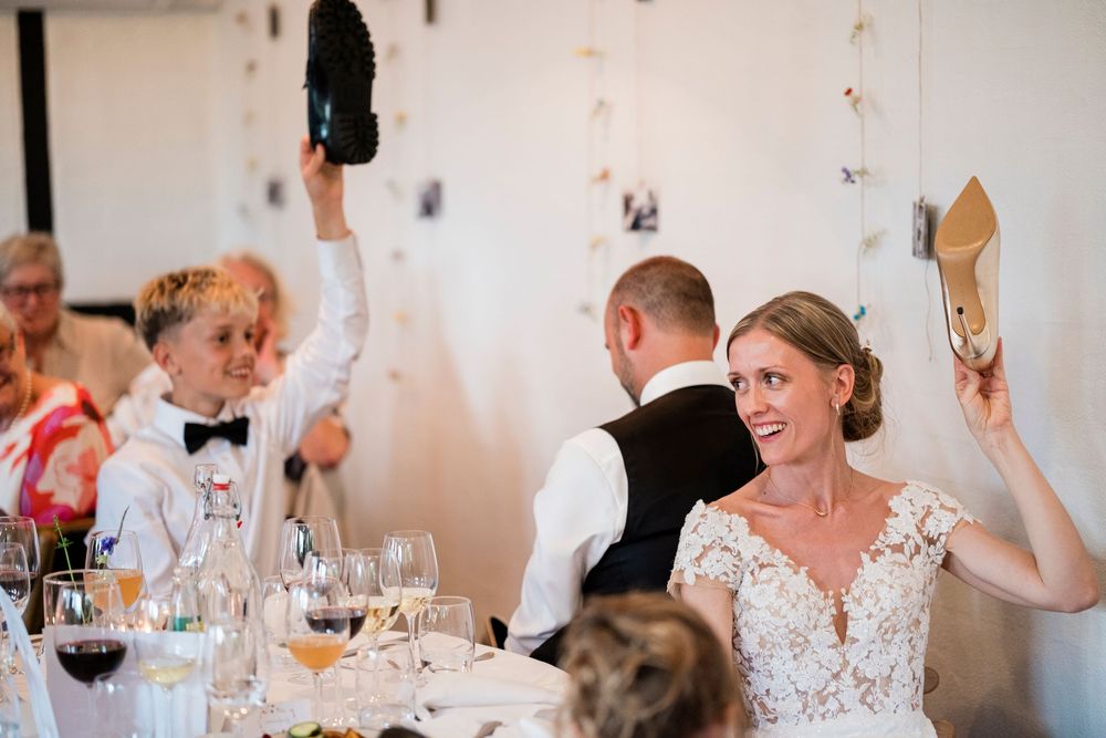 Bride in lace wedding dress laughs while holding shoe up during fun wedding reception game.