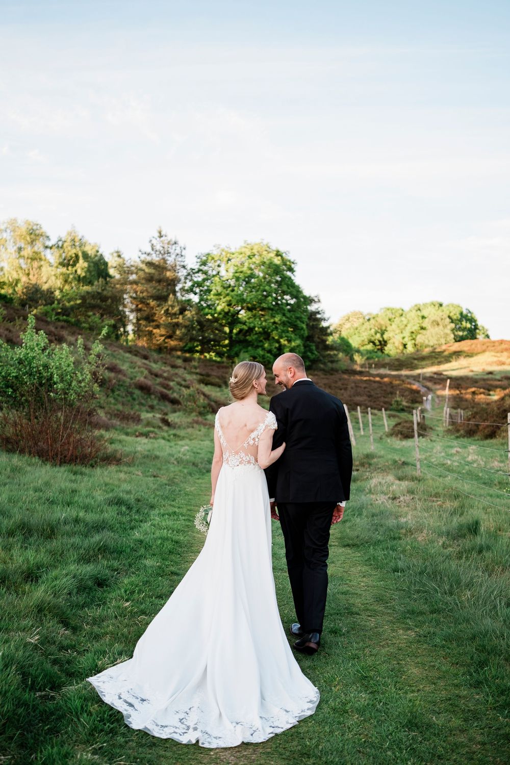 Bride and groom walking hand in hand through lush green countryside, bride wearing elegant white gown with long train.