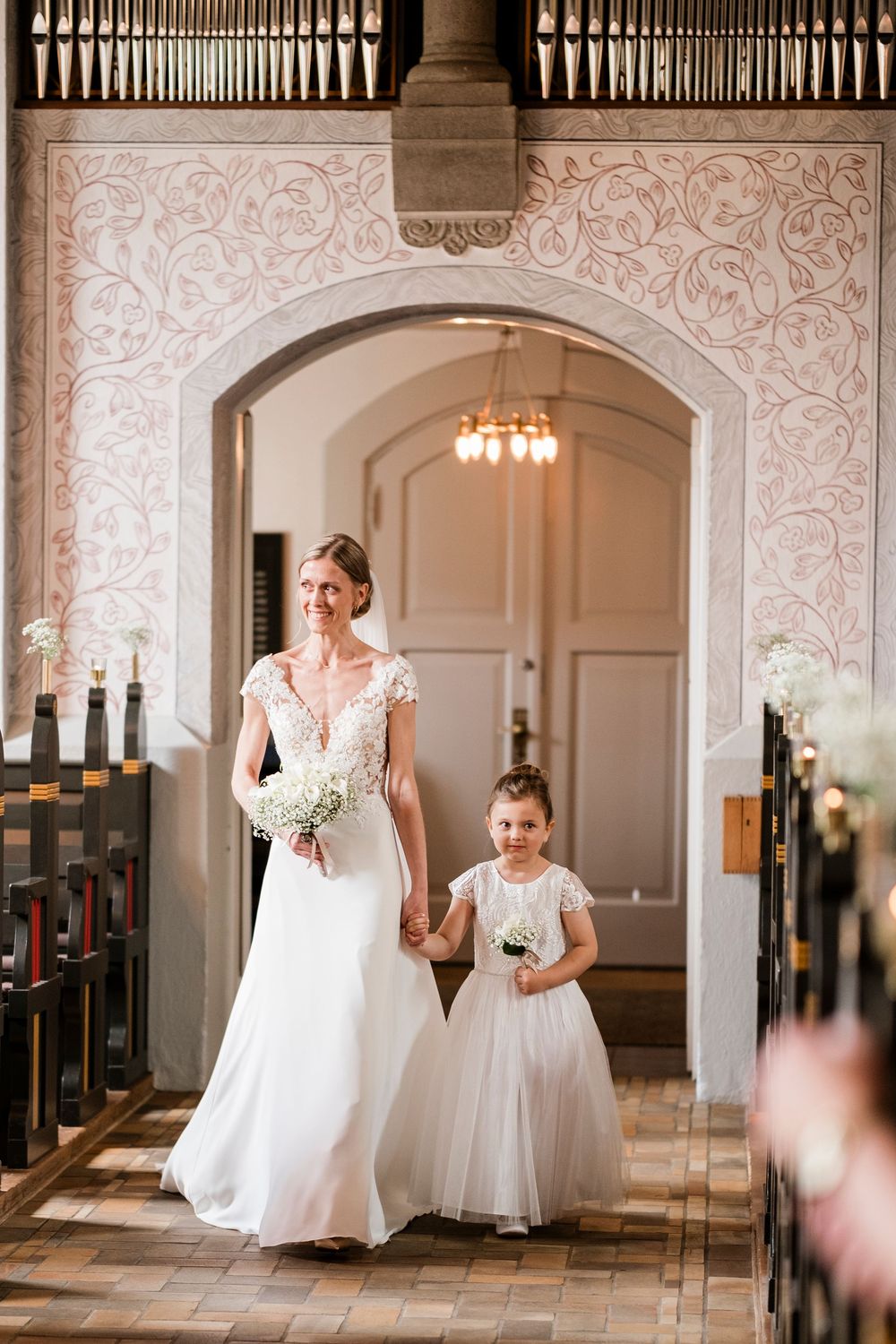 Bride in white lace gown walks down church aisle holding hands with young flower girl in white dress.