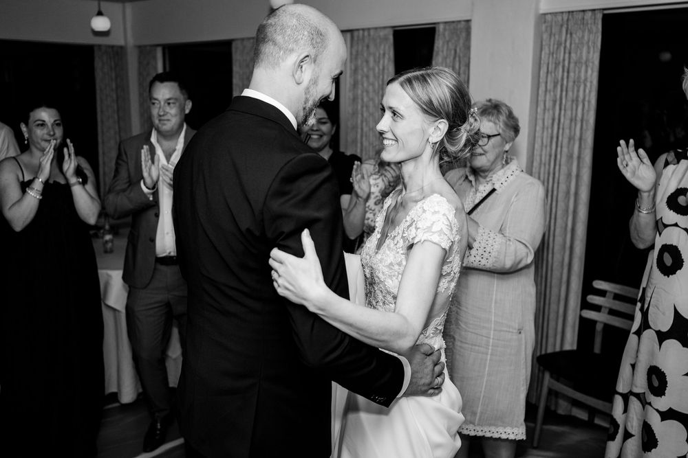 Bride and groom share first dance at wedding reception in black and white photo with guests clapping.