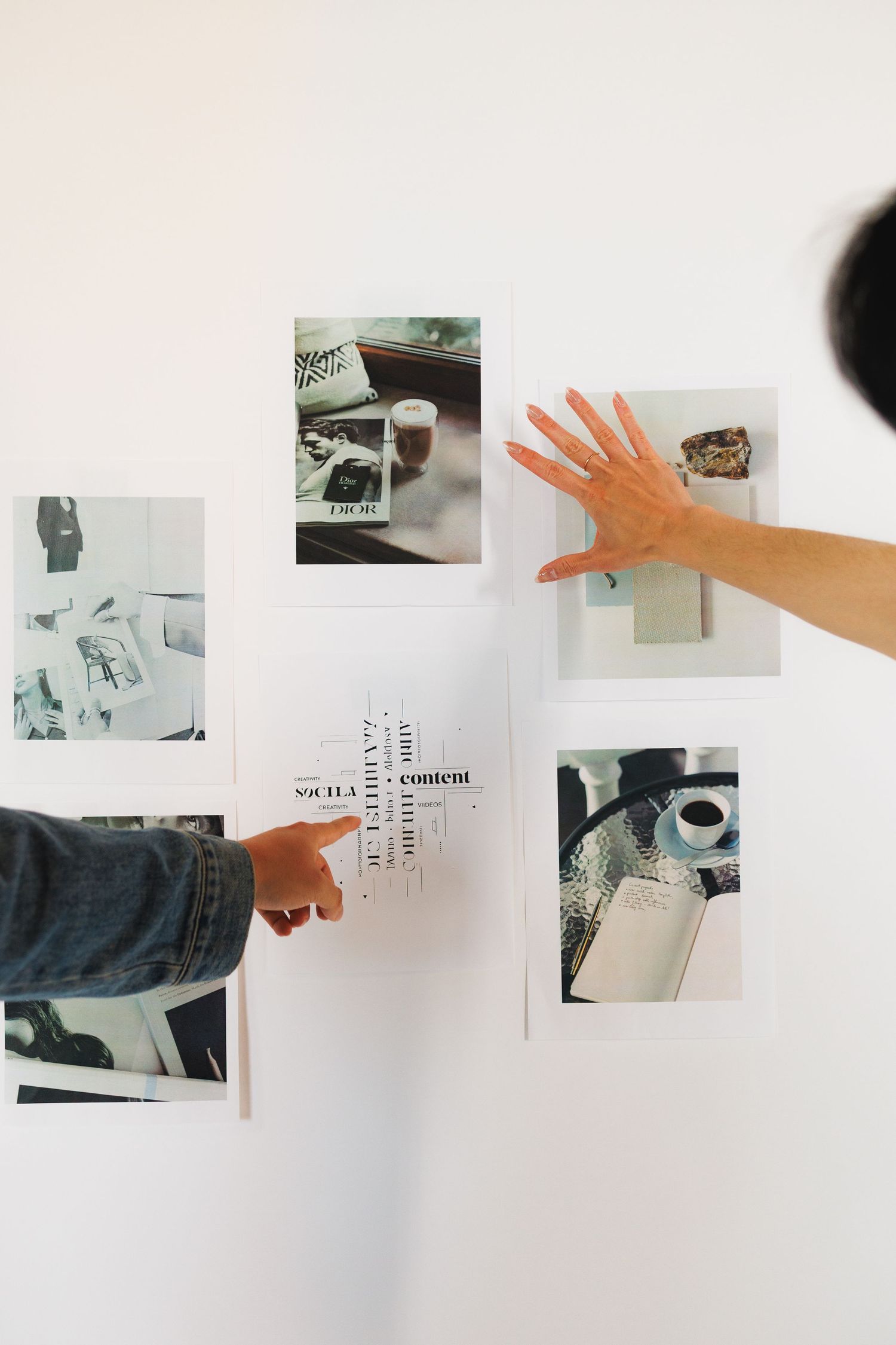 Two people arranging printed photos and typography cards on a white wall in a creative mood board layout.