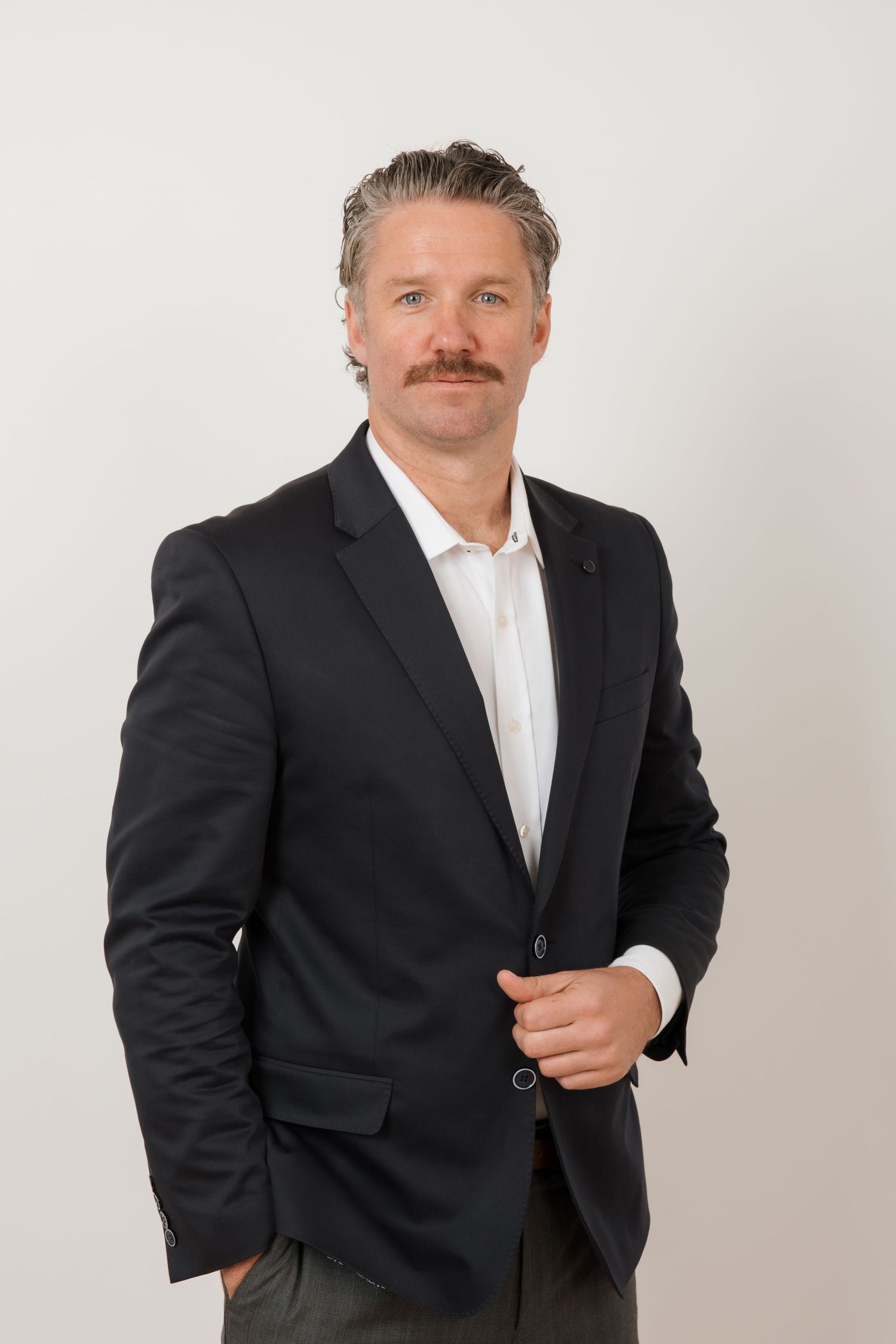 Smiling man in white Irish Mini t-shirt and black cap sits on white stool against blue background.