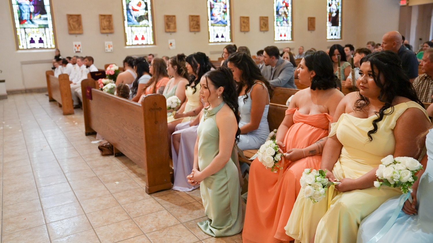 Bridesmaids in pastel gowns seated in church pews during a wedding ceremony with stained glass windows.