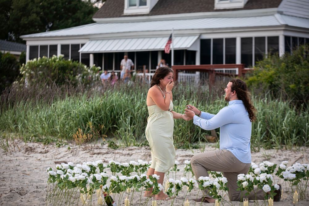 Man proposes to woman on beach with white flower arrangement, house in background at sunset.