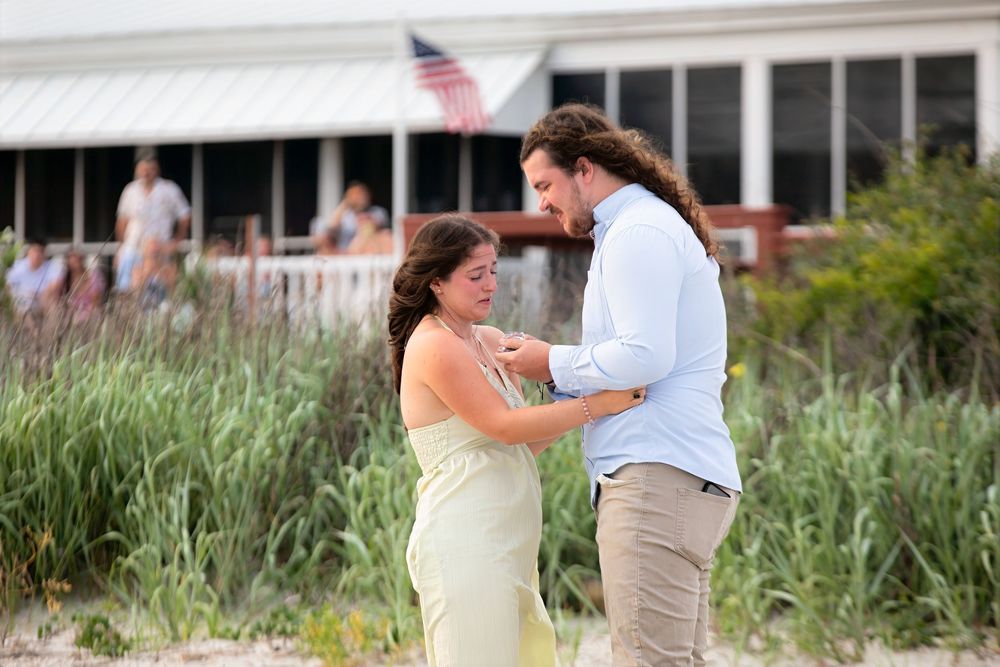 Couple sharing a romantic moment on a sandy beach near tall grass, with an American flag and building in background.