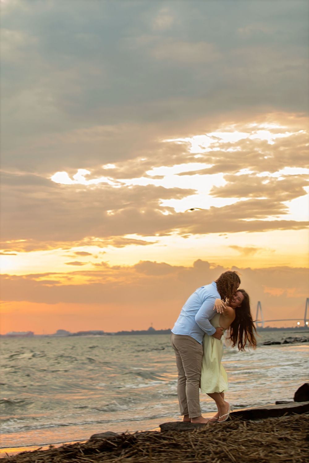 Couple sharing a romantic kiss on a rocky beach at sunset with dramatic orange and golden cloudy sky backdrop.