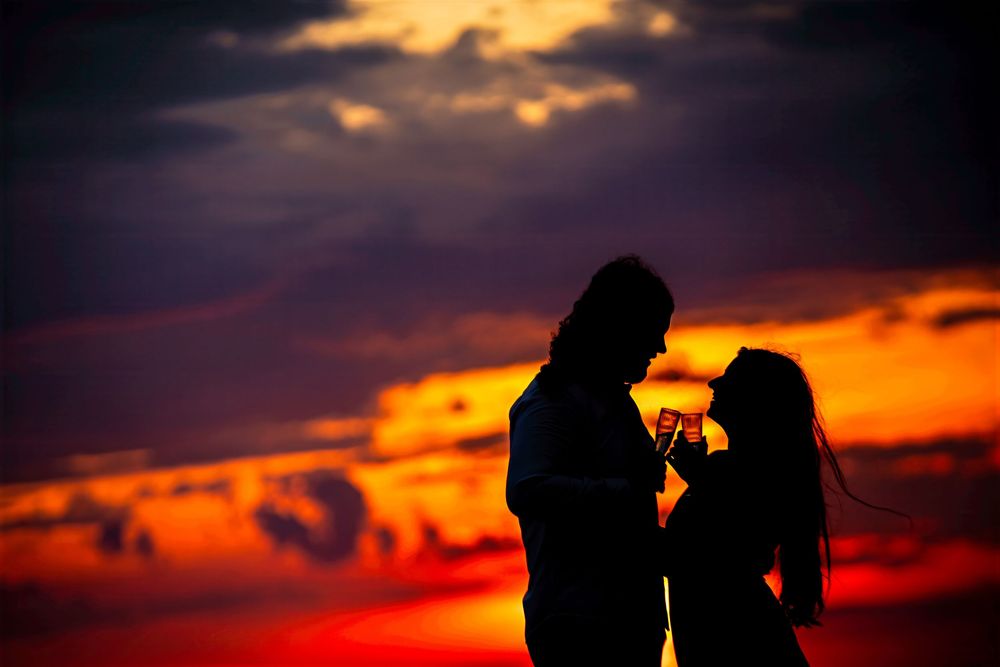 Romantic couple silhouette holding hands against a vivid red and orange sunset sky with dramatic clouds.