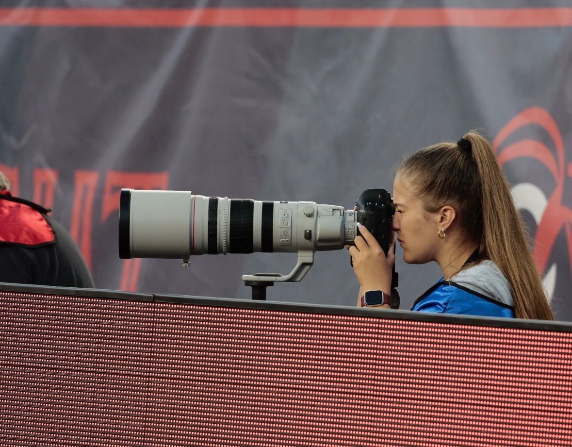 Young female photographer using a large telephoto lens camera at a sporting event.