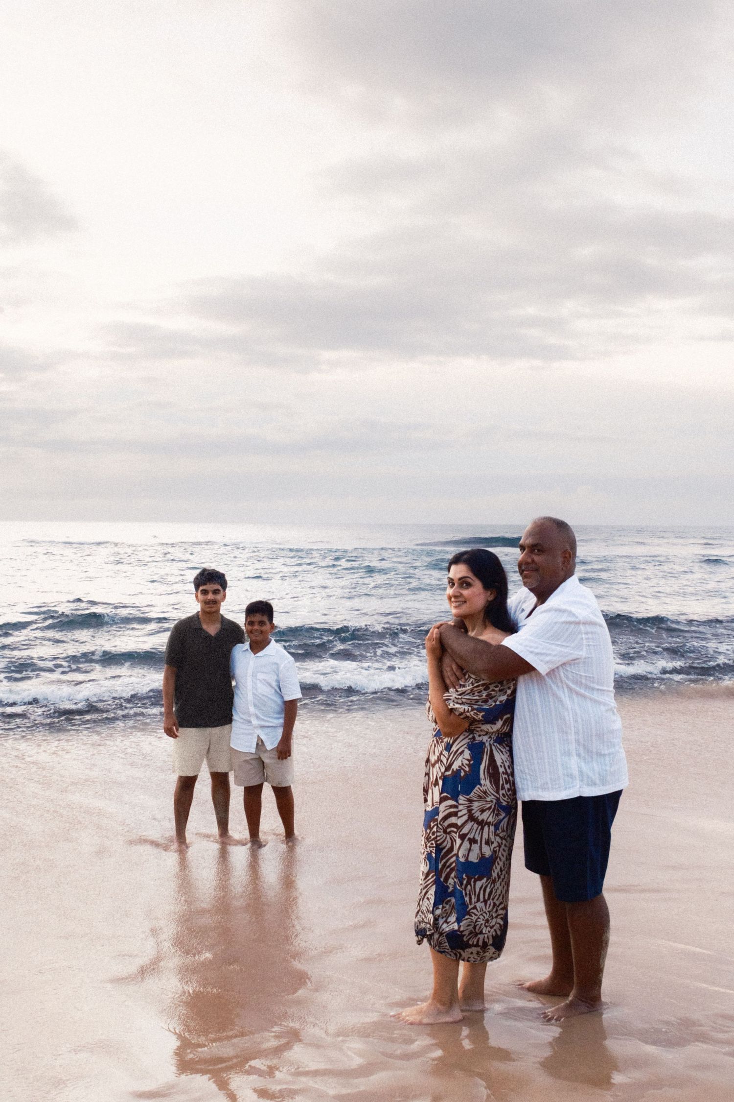 Family of four enjoying a beach vacation, posing on wet sand with ocean waves in the background at dusk.