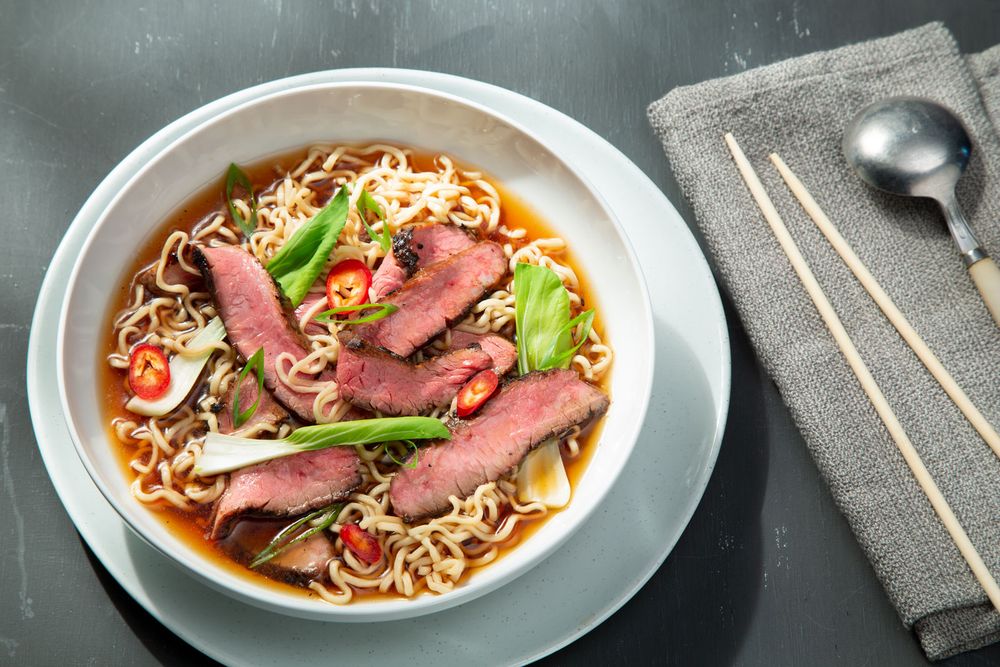 Sliced grilled beef steak ramen noodle soup with chili peppers in a white bowl.