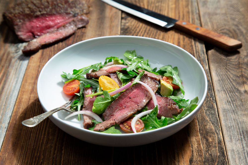 Sliced medium-rare steak salad with cherry tomatoes and arugula in a white bowl.