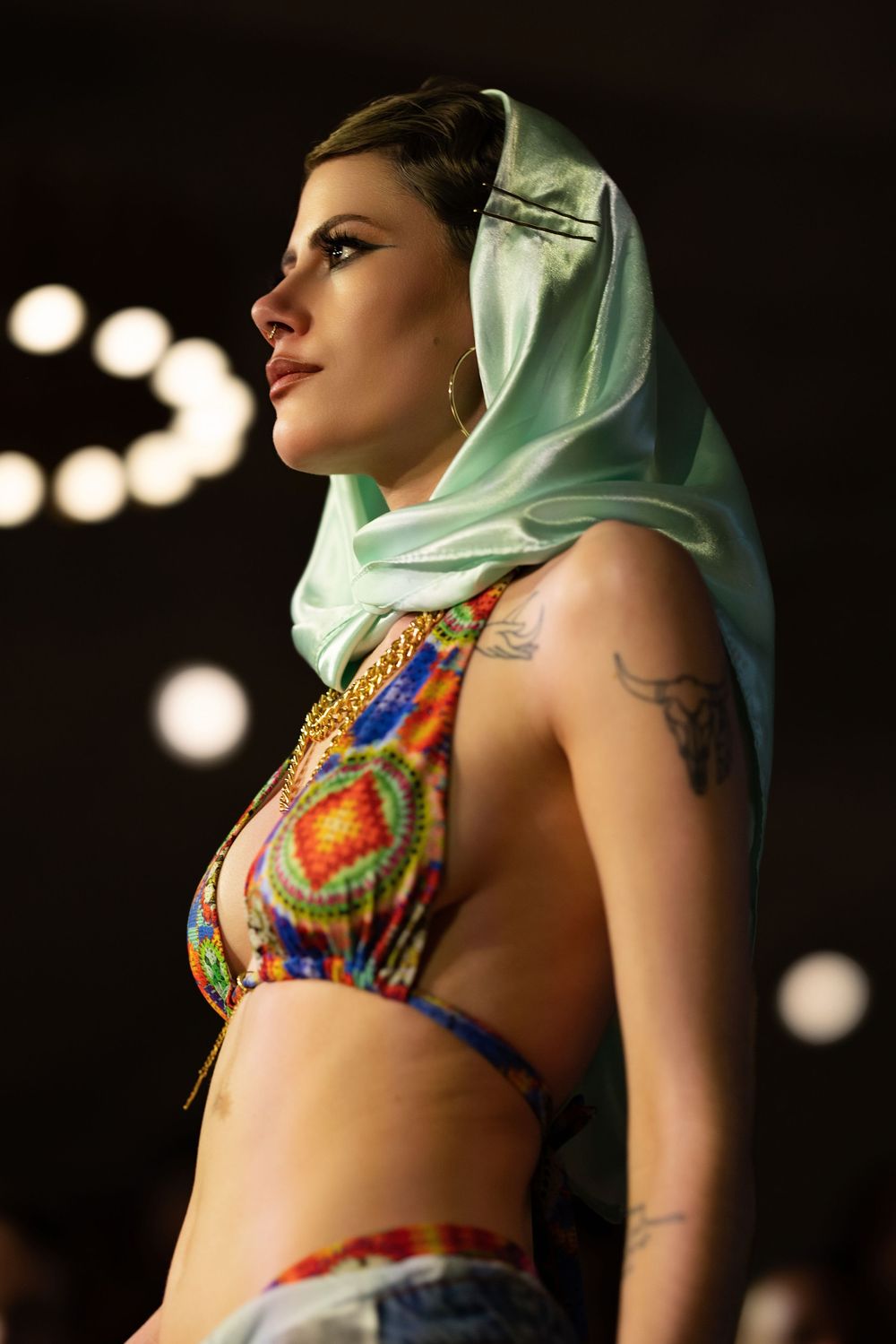 Model wearing colorful printed crop top with mint green headscarf and gold jewelry on runway.