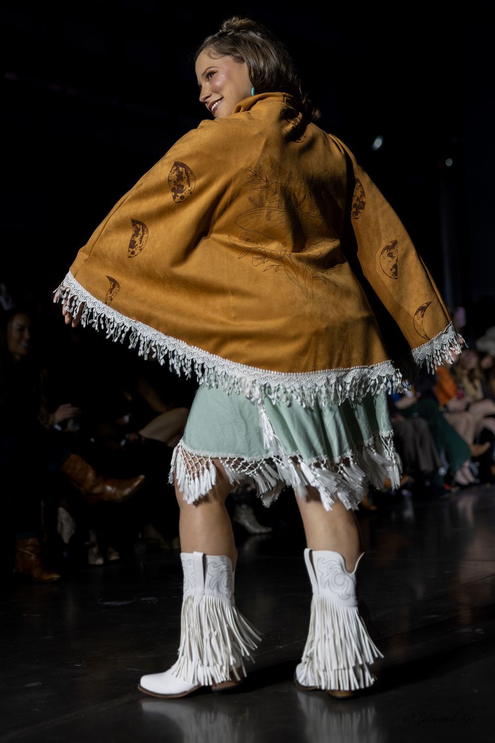 Model wearing mustard suede fringe jacket with mint skirt and white fringe boots on runway.