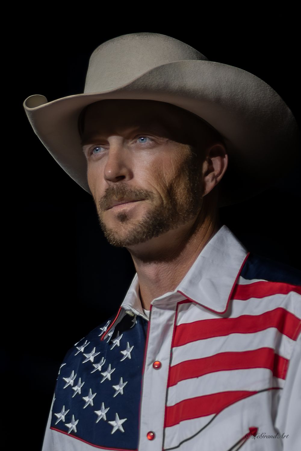 Rugged cowboy in American flag shirt and white Stetson hat poses against dark background.