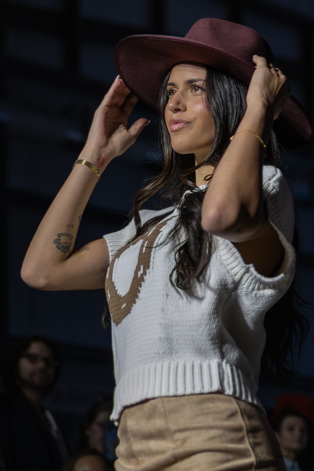 Stylish woman in white sweater adjusts burgundy wide-brim hat at fashion event.