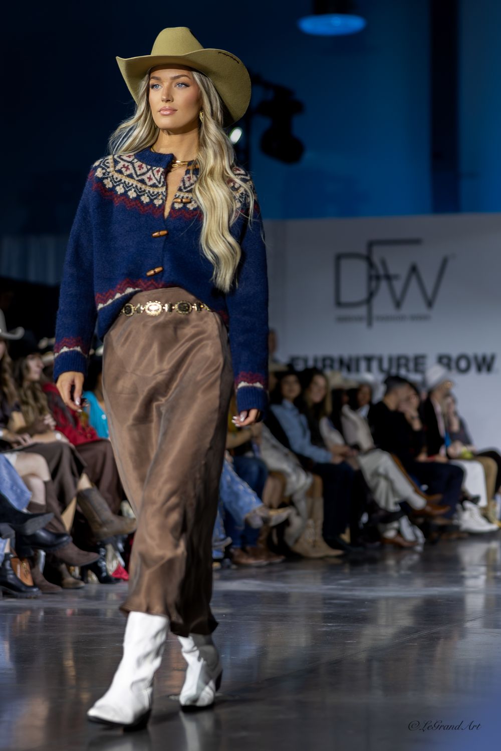 Blonde model walks runway in western-style navy sweater, brown trousers, and white boots.