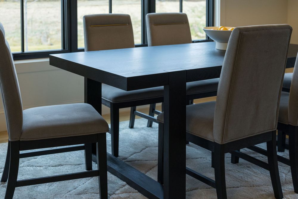 Dark wood rectangular dining table with upholstered chairs on area rug near large windows.