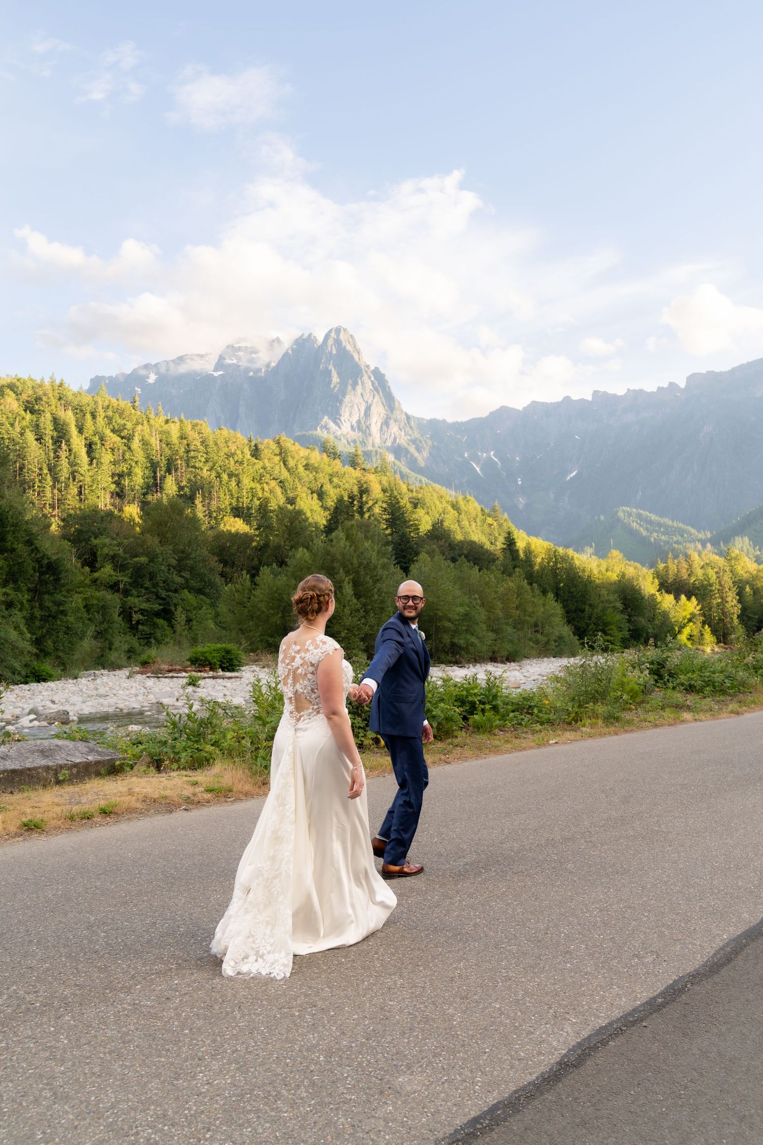 Bride and groom hold hands on mountain road with scenic alpine forest and peaks backdrop on wedding day.