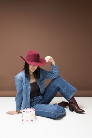 Woman in denim outfit and burgundy cowboy hat sitting on floor beside a small decorated cake.