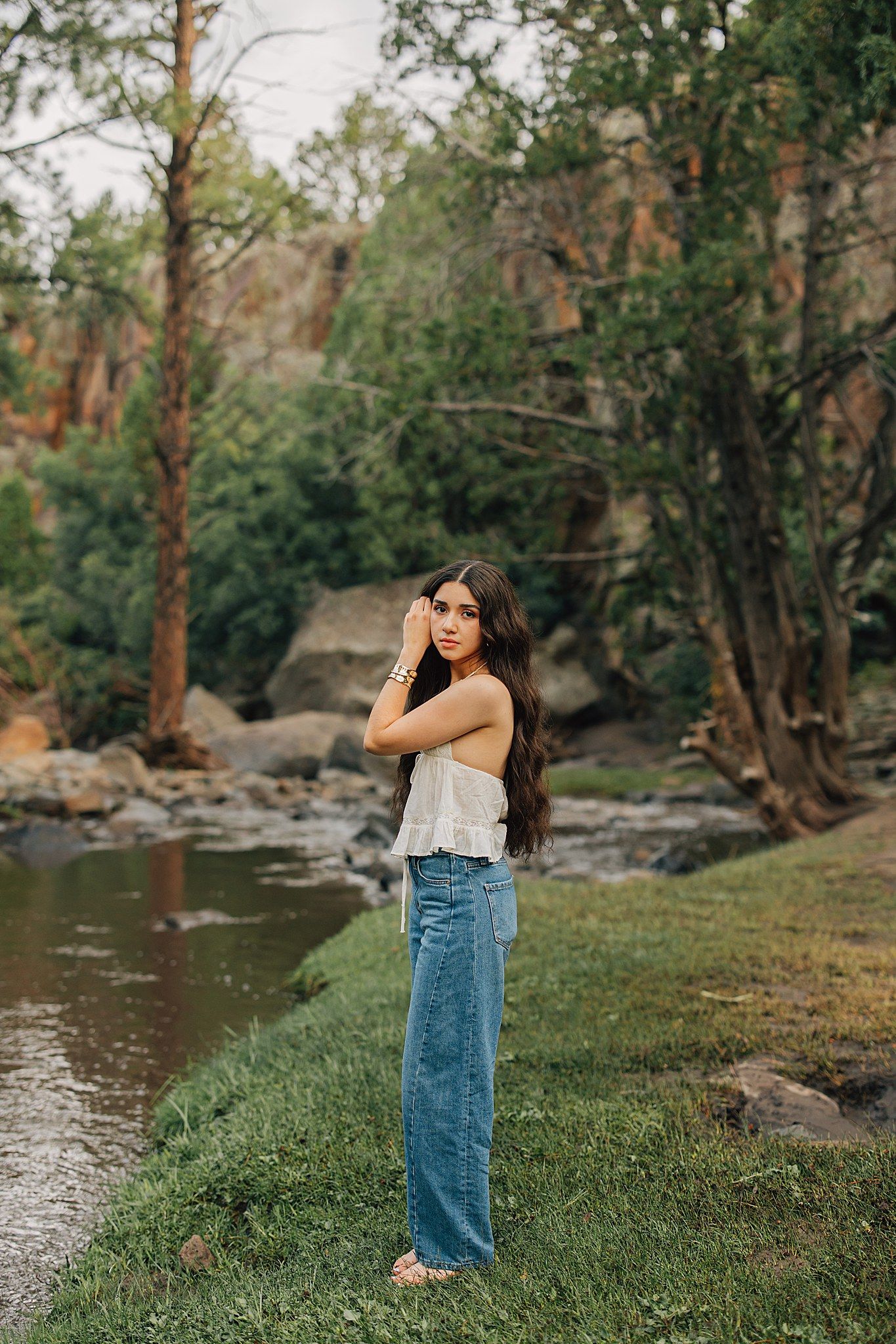 Young woman in white top and wide-leg jeans poses by a rocky creek surrounded by lush green trees.