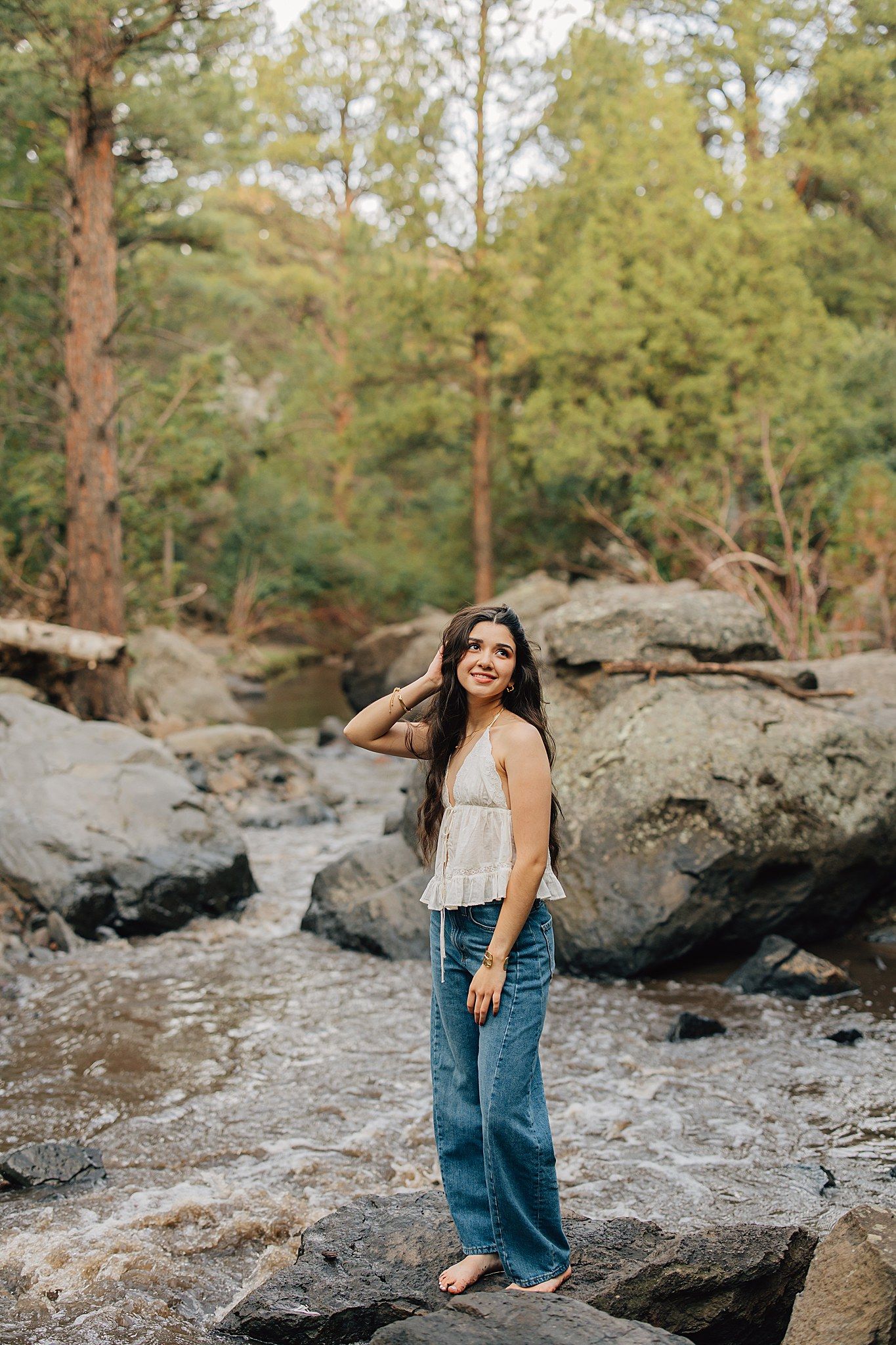 Young woman in wide-leg jeans and white top standing on rocks by a forest stream, smiling outdoors.