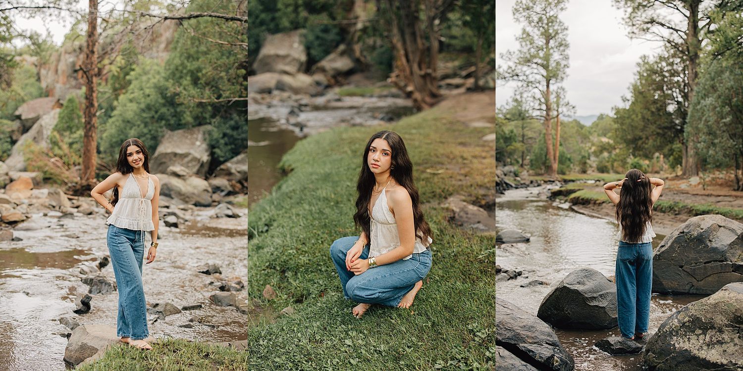 Young woman in jeans and tank top poses near a rocky creek surrounded by lush green trees in a nature setting.