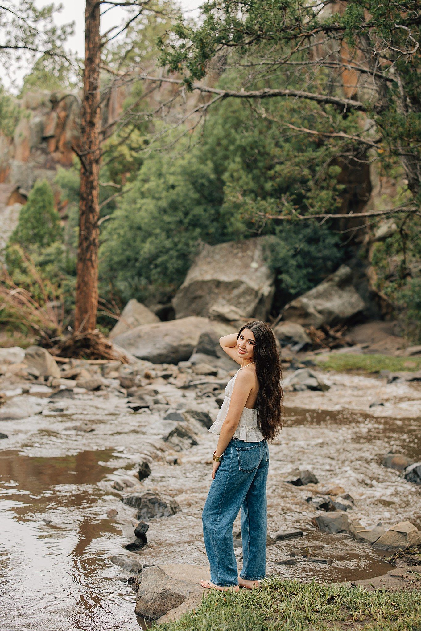 Young woman in white top and blue jeans smiling by a rocky creek surrounded by lush green trees.