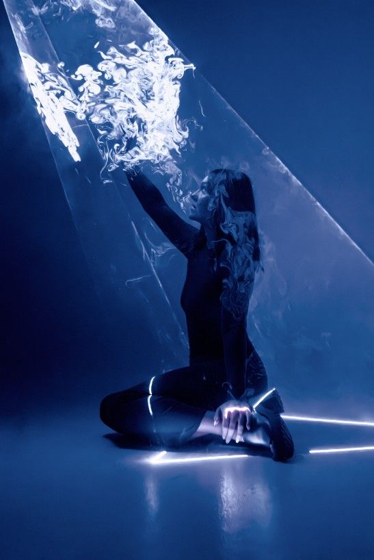 Person in blue wig and dark outfit sits on floor under neon purple and yellow studio lights.