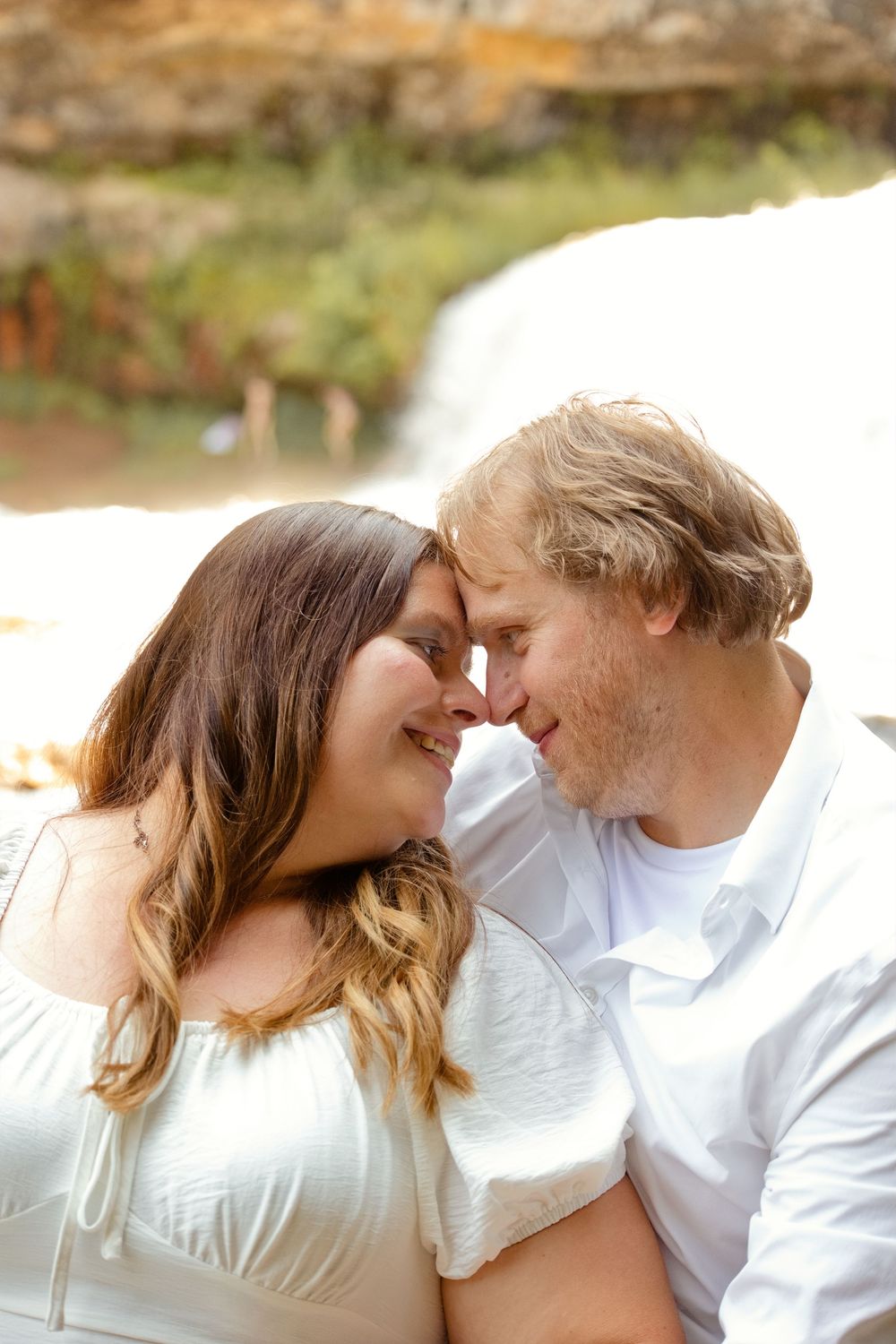 Romantic couple touching foreheads tenderly in front of a waterfall, wearing white outfits in a loving embrace.