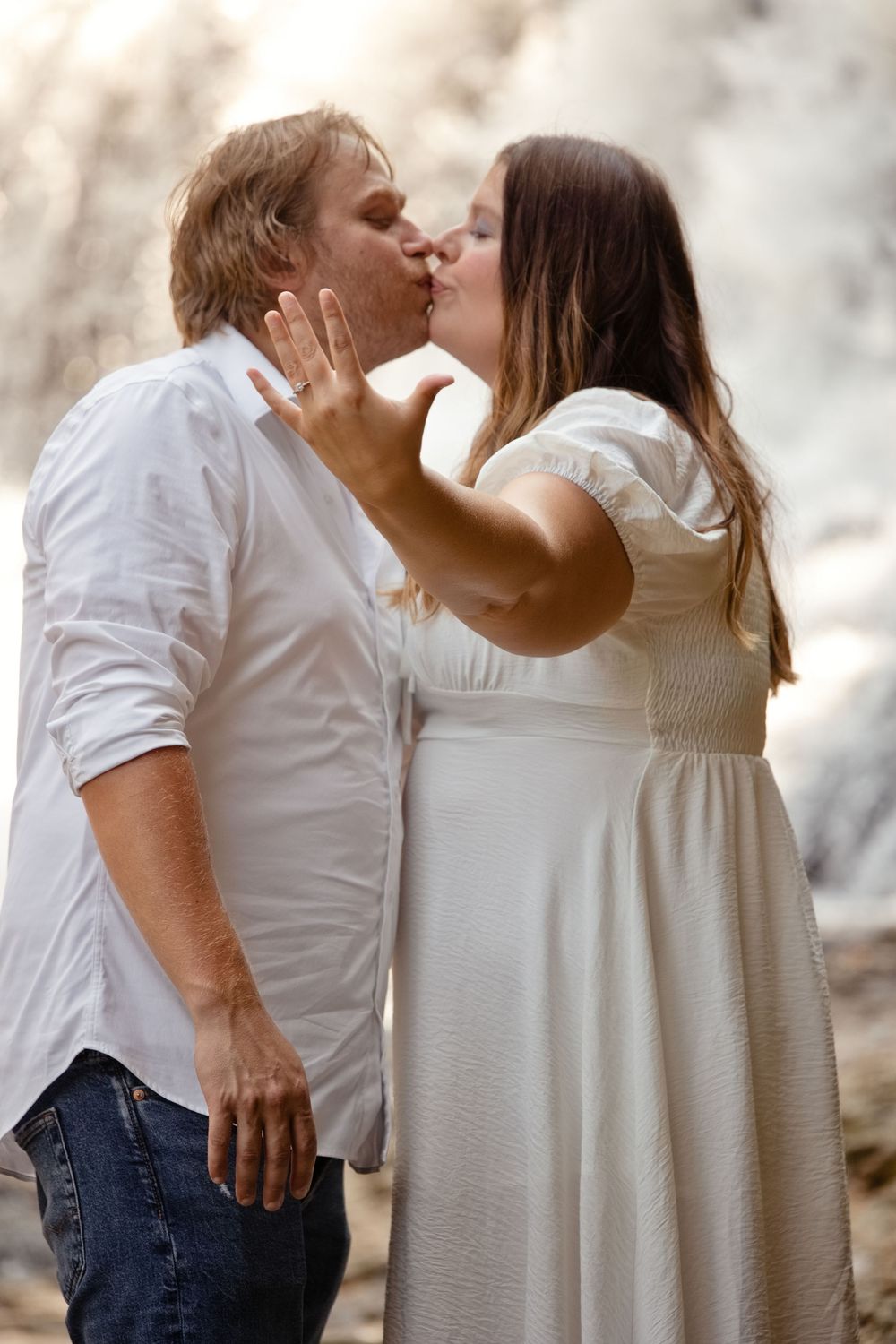 Couple kissing outdoors, man in white shirt and jeans, woman in white dress showing engagement ring.