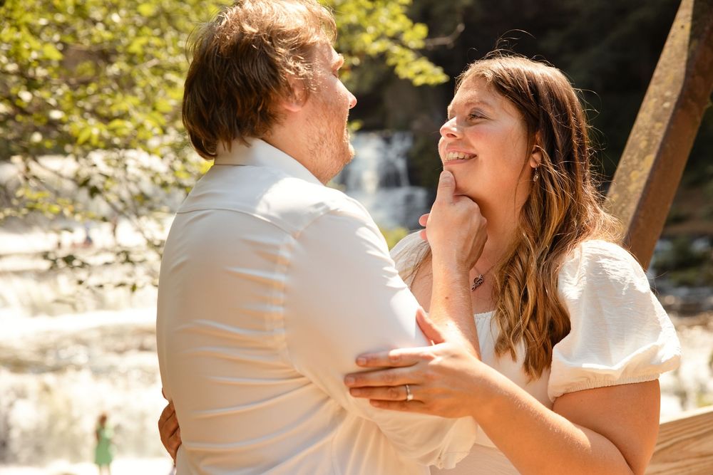 Couple embracing and smiling at each other outdoors near a waterfall, dressed in white for a romantic occasion.
