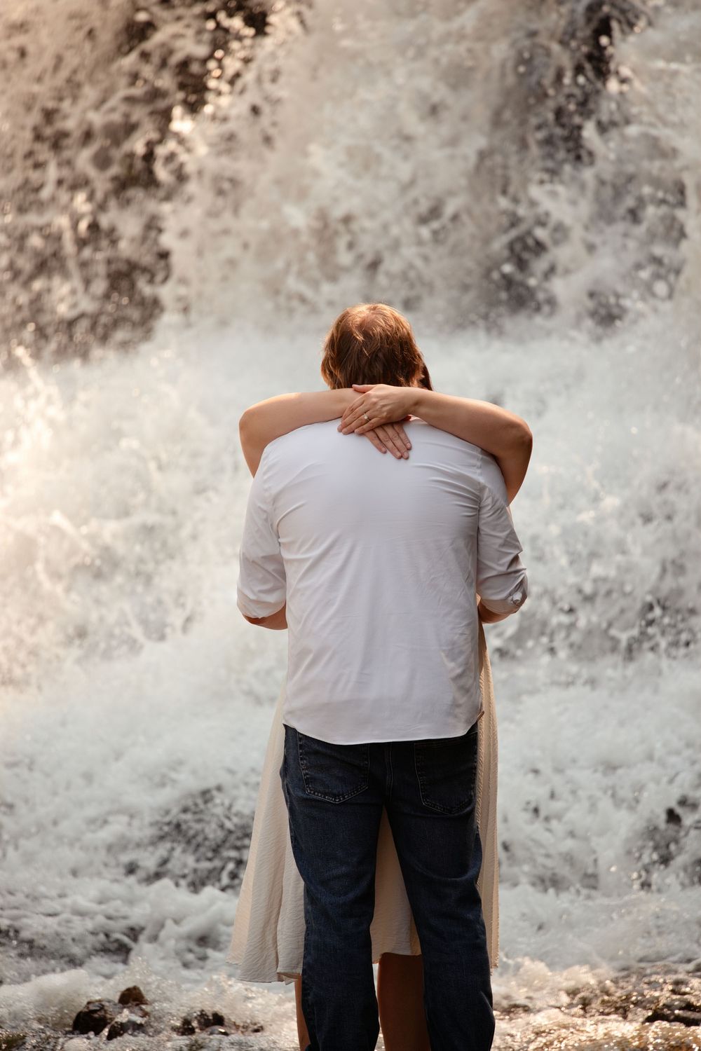 Couple embracing romantically in front of a rushing waterfall, viewed from behind.