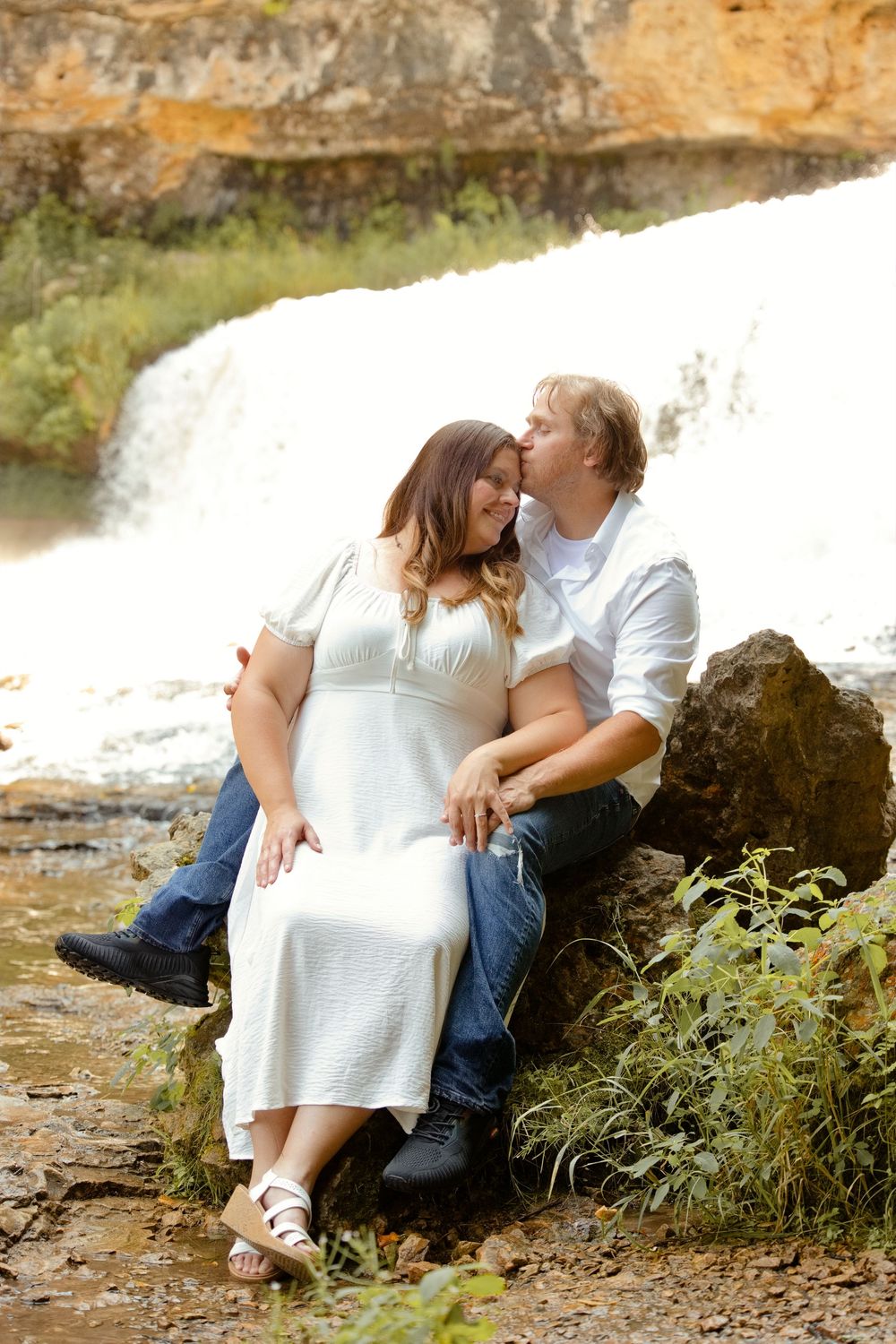 Couple sitting on rocks by a waterfall, man kissing woman's cheek, woman in white dress, romantic outdoor portrait.