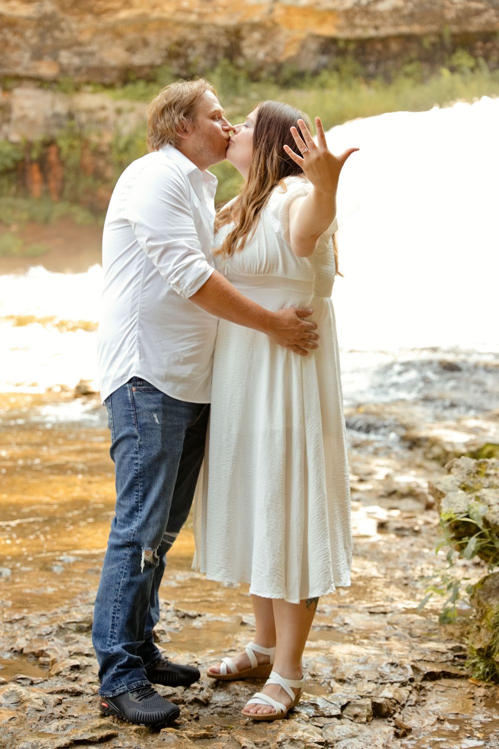 Couple sharing a romantic kiss outdoors near a waterfall, man in white shirt and jeans, woman in white dress.