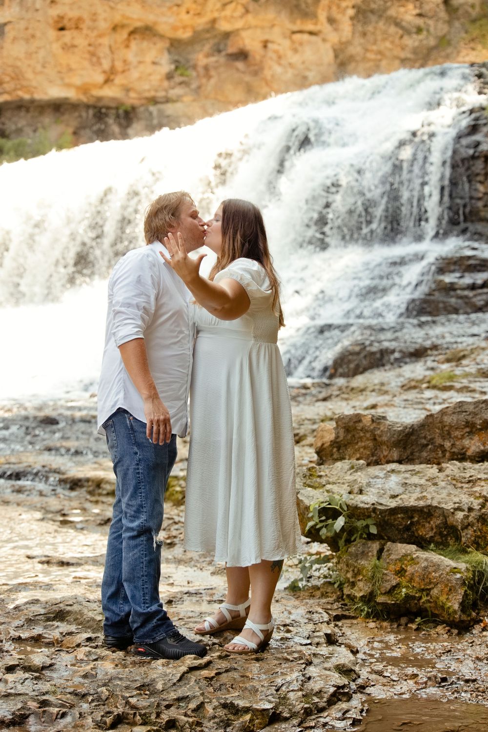 Couple kissing romantically in front of a cascading waterfall, woman in white dress, man in white shirt and jeans.