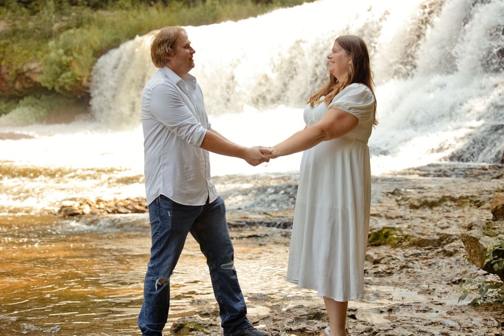 Couple holding hands and smiling by a waterfall, standing in shallow water on rocky shore.