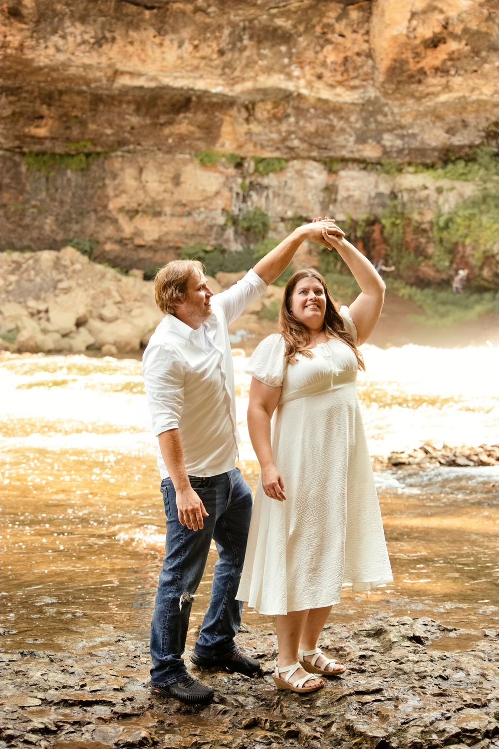Couple dancing outdoors near a rocky waterfall, man in white shirt and jeans spinning woman in white dress on wet stones.