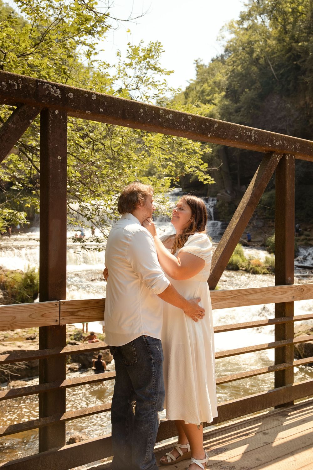 Couple embracing on a wooden bridge with a waterfall and lush green trees in the background on a sunny day.