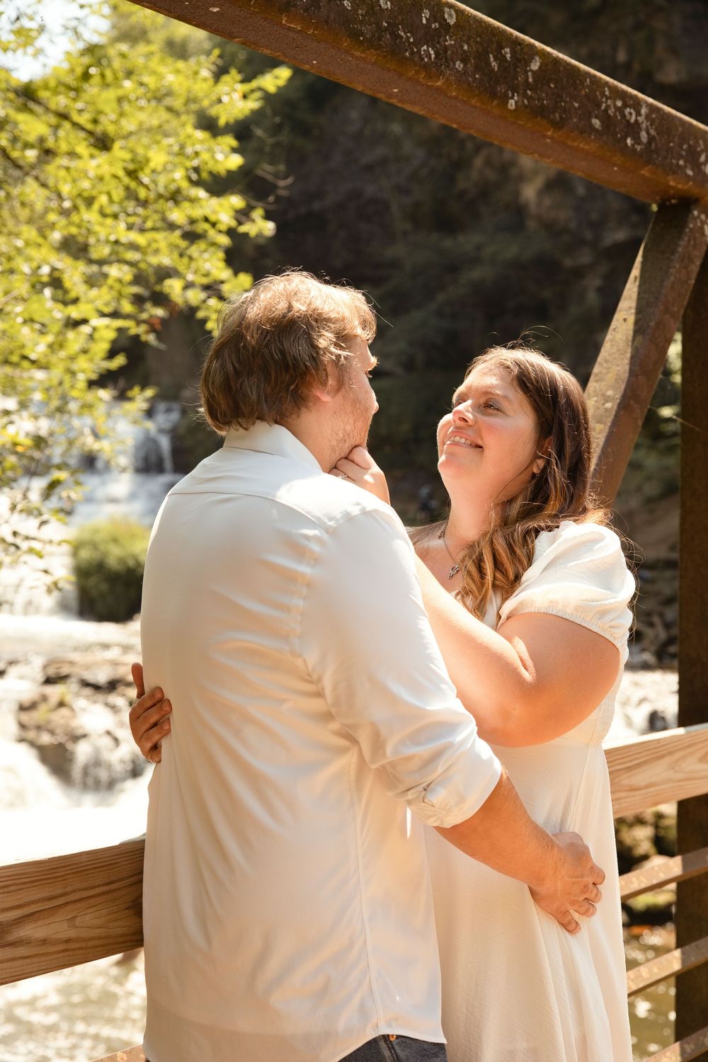 Couple in white attire embracing on a wooden bridge near a waterfall, sharing a romantic moment outdoors.