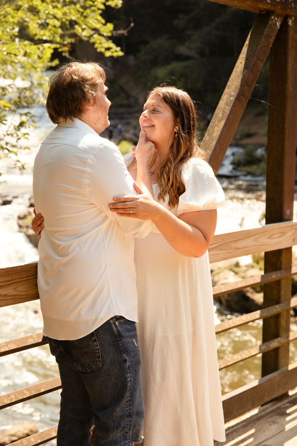 Happy couple in white outfits holding each other on a rustic wooden bridge with a scenic waterfall backdrop.