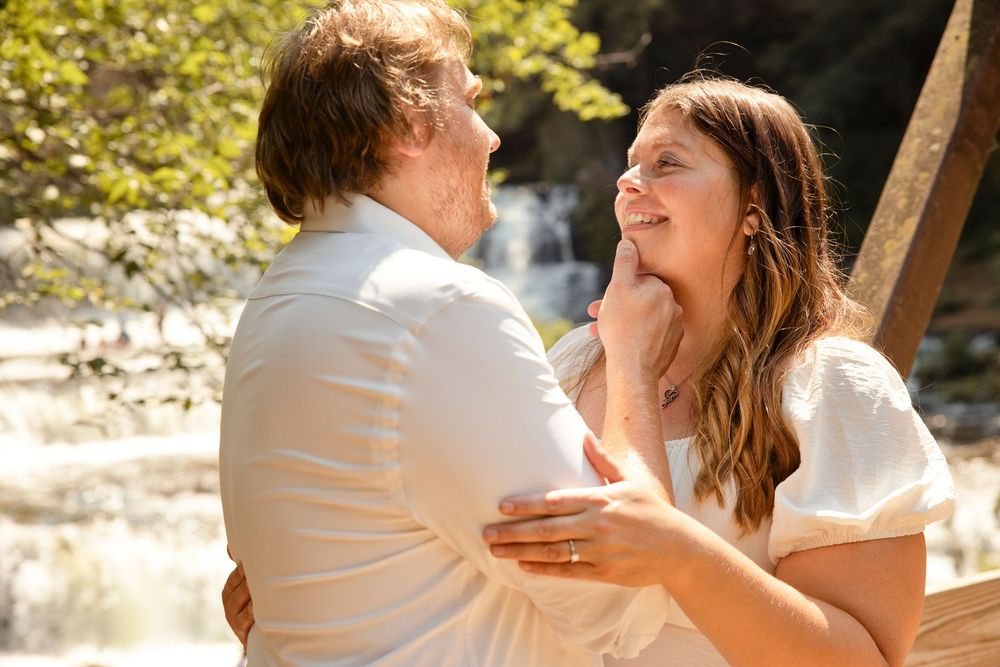 Couple embracing and smiling at each other outdoors near a waterfall, dressed in white for a romantic occasion.